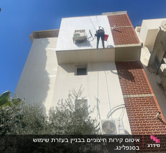 אדם עובד בגובה על קיר בניין עם חבלי בטיחות